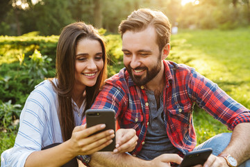 Image of beautiful couple man and woman using cellphones while resting in green park