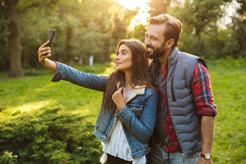 Image of cute couple man and woman taking selfie photo on cellphone while walking in green park