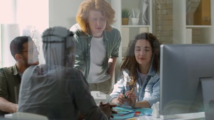 Panning medium shot, through glass pane, of 20-something red-haired creative designer standing in brightly lit studio and showing presentation to his multiethnic colleagues on computer display