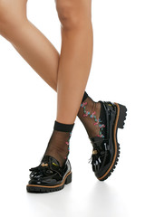 Cropped side shot of woman's legs in black nylon socks, adorned with multicolored floral needlework. The girl in patent leather shoes with ribbed soles and tassels, is posing on white background.