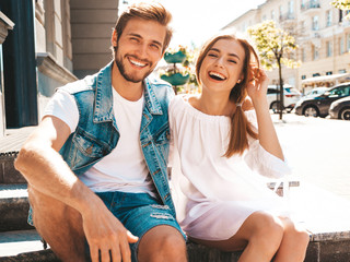 Smiling beautiful girl and her handsome boyfriend. Woman in casual summer dress and man in jeans clothes. Happy cheerful family. Sitting on stairs on the street background.Hugging couple