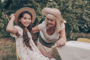 Family portrait of a happy young granddaughter dressed in a sundress and hat and an elderly grandmother on a summer day in the park. Beautiful girl poses for the camera 