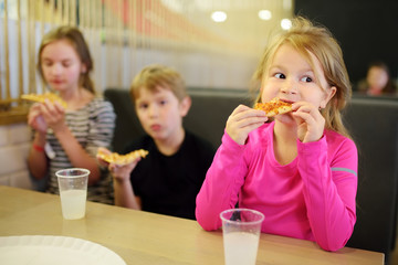 Three cute funny siblings eating slices of pizza at indoor restaurant or cafe.