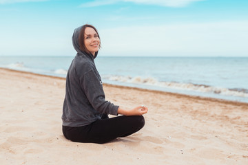 Fototapeta premium A woman smiles and practices meditation on the seashore, the ocean. Practice clearing thoughts, calming down. Yoga on the coast.