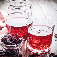 Closeup view at glass of hibiscus ice tea and jug on wooden table background