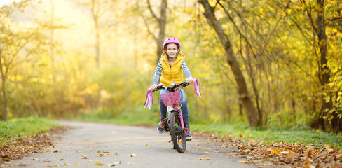 Obraz premium Adorable young girl riding a bike in a city park on sunny autumn day. Active family leisure with kids.
