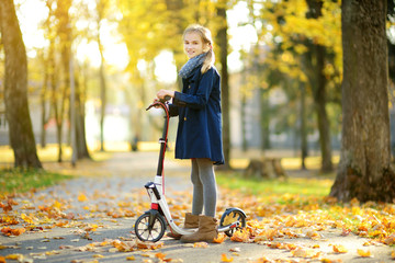 Adorable young girl riding her scooter in a city park on sunny autumn evening. Pretty preteen child riding a roller.