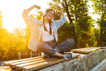 Pleased happy young teenage girl student sitting outdoors in beautiful green park listening music with headphones.