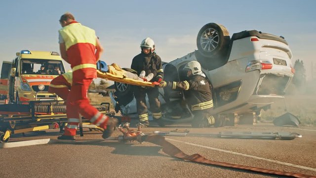 On the Car Crash Traffic Accident Scene: Paramedics Saving Life of an Accident Victim who is Lying on Stretchers. They Listen To a Heartbeat and Give First Aid. Firefighters Running Extinguishing Fire