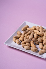 Peanuts in white square shaped plate minimal on purple background backdrop food nut texture copy space angle view