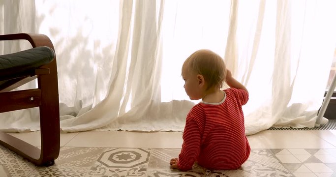 Back view of unrecognizable toddler in red striped romper sitting on floor and looking at window with curtains at home