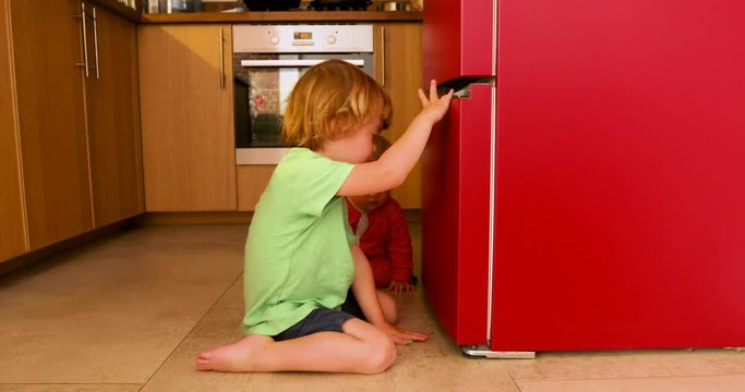 Side view of cute children sitting and playing on floor in kitchen near fridge