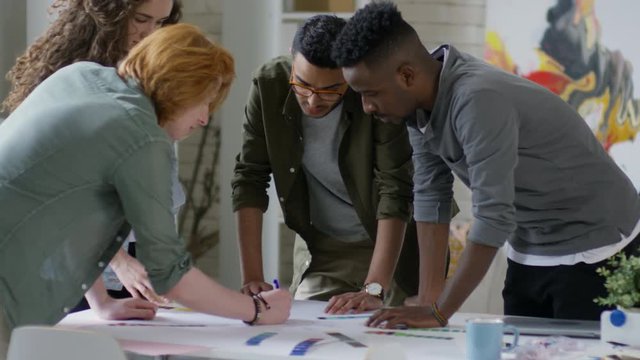 Medium tilting shot of four multiethnic creative designers in their twenties, standing around table and drawing flowchart of new design project, proposing colour combinations for different parts