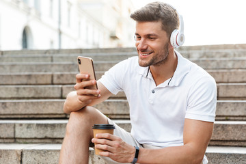 Portrait of masculine young man using headphones and smartphone while sitting on stairs outdoors with paper cup