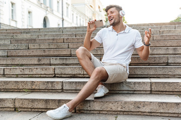 Portrait of happy young man wearing headphones holding smartphone and singing outdoors