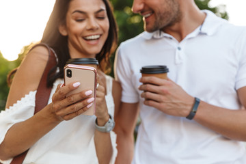 Portrait of happy young couple using smartphone while walking outdoors with paper cups