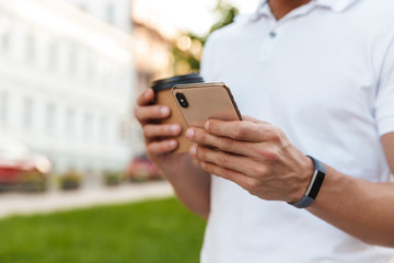 Cropped portrait of caucasian young man holding paper cup and smartphone while walking outdoors