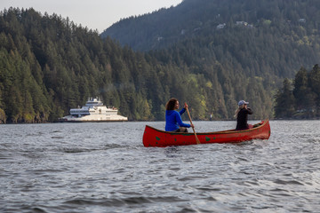 Adventurous friends on a red canoe are paddling in the Howe Sound with Ferry Boat in the Background during a cloudy day. Taken near Bowen Island, West of Vancouver, BC, Canada.