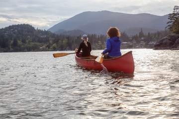 Couple adventurous female friends on a red canoe are paddling in the Howe Sound during a cloudy sunset. Taken near Bowen Island, West of Vancouver, BC, Canada.