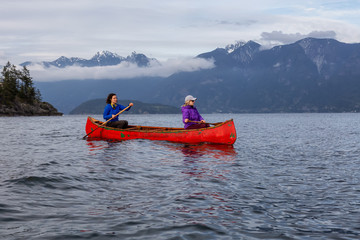Couple adventurous female friends on a red canoe are paddling in the Howe Sound during a cloudy...