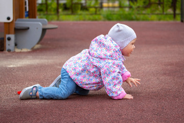 Baby girl crawling on the playground. A child in warm clothes on all fours. A 1 year old baby walks near the house in spring autumn clothes.