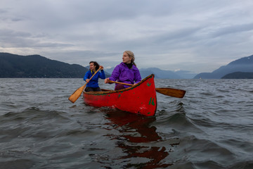 Couple adventurous female friends on a red canoe are paddling in the Howe Sound during a cloudy sunset. Taken in Horseshoe Bay, West Vancouver, BC, Canada.