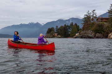 Couple adventurous female friends on a red canoe are paddling in the Howe Sound during a cloudy sunset. Taken in Horseshoe Bay, West Vancouver, BC, Canada.