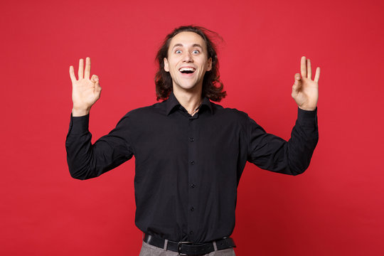 Stylish young curly long haired man in black shirt posing isolated on red wall background studio portrait. People sincere emotions lifestyle concept. Mock up copy space. Hold hands in yoga gesture,