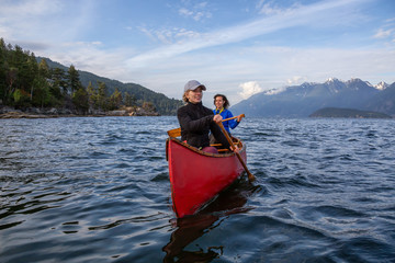 Obraz premium Couple adventurous female friends on a red canoe are paddling in the Howe Sound during a cloudy and sunny evening. Taken near Bowen Island, West of Vancouver, BC, Canada.