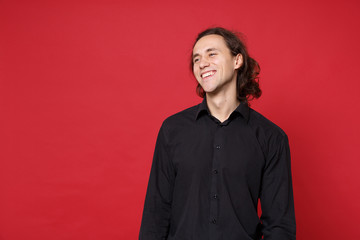 Handsome stylish young curly long haired man in black shirt posing isolated on red wall background studio portrait. People sincere emotions lifestyle concept. Mock up copy space. Looking aside smile.