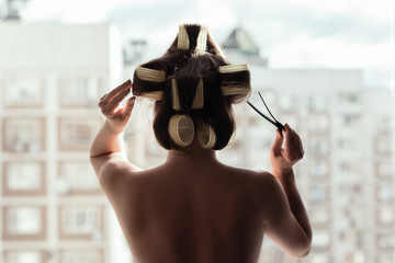 naked young woman in a huge hair curlers standing near window opposite high building