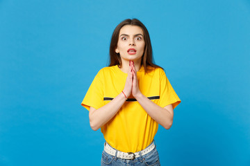 Portrait of shocked young woman in vivid casual clothes looking camera, holding hands folded, praying isolated on bright blue wall background in studio. People lifestyle concept. Mock up copy space.