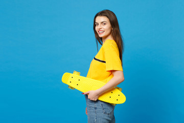 Side view of smiling young woman in vivid casual clothes standing, looking camera, holding yellow skateboard isolated on blue wall background in studio. People lifestyle concept. Mock up copy space.