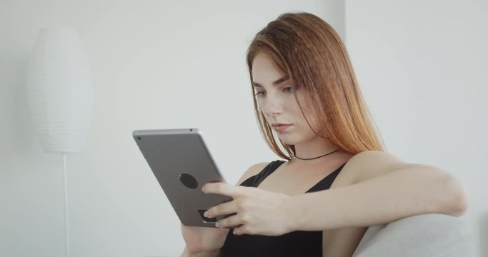 Closeup shoot of young sexy caucasian female in black bodysuit using the tablet looking at camera smiling happily sitting on the couch indoors