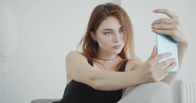 Closeup shoot of young sexy caucasian female in black bodysuit taking selfies on the phone posing sitting on the couch indoors