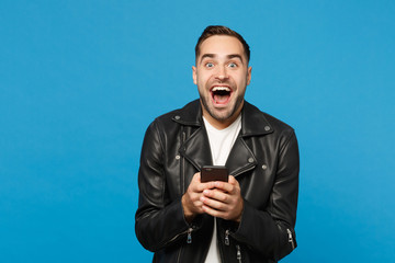 Handsome fun overjoyed excited young unshaven man in black leather jacket white t-shirt using mobile phone isolated on blue wall background studio portrait. People lifestyle concept Mock up copy space