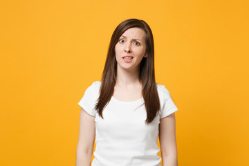 Portrait of perplexed puzzled young woman in white casual clothes standing and looking camera isolated on bright yellow orange wall background in studio. People lifestyle concept. Mock up copy space.