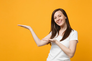 Portrait of smiling pleasant young woman in white casual clothes looking camera, pointing hands aside isolated on yellow orange wall background in studio. People lifestyle concept. Mock up copy space.