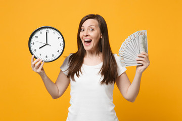 Portrait of cheerful young woman in white casual clothes holding round clock, fan of cash money in dollar banknotes isolated on yellow orange background. People lifestyle concept. Mock up copy space.
