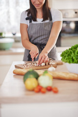Beautiful smiling dedicated Caucasian brunette in apron standing in kitchen and chopping mushrooms. On table are lots of vegetables. Cooking at home concept.
