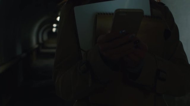Close-up shot of businesswoman in a coat going through the tunnel in the evening, getting out of pocket mobile phone, reading, smiling and texting back