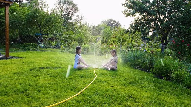 two little girls sitting on green lawn near working garden sprinkler
