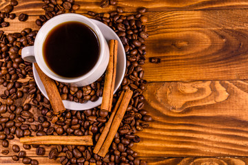 Cup of hot coffee, cinnamon sticks and scattered coffee beans on wooden table. Top view