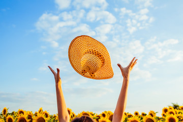 Young woman walking in blooming sunflower field throwing hat up and having fun. Summer vacation