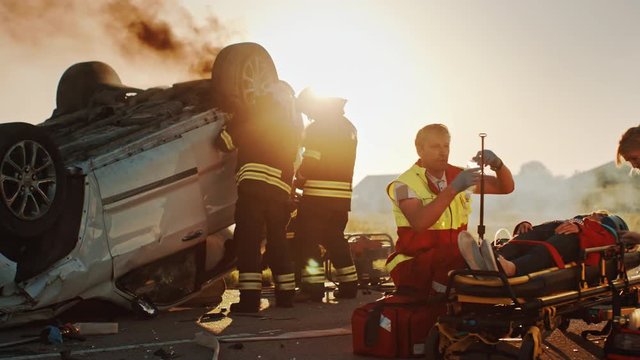On the Car Crash Traffic Accident Scene: Paramedics Save Life of a Passenger Lying on Stretchers. They Listen To a Heartbeat, Use Oxygen Mask, setup Drop Counter. Firefighters Use Cutters to Open Car