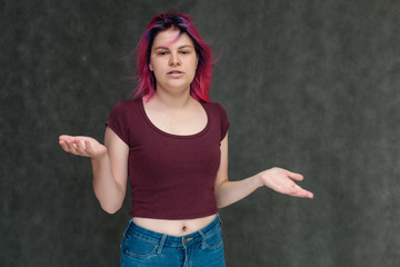 Portrait to the waist of a young pretty teen girl in a burgundy T-shirt and jeans with beautiful purple hair on a gray background in the studio. Talking, smiling, showing hands with emotions.
