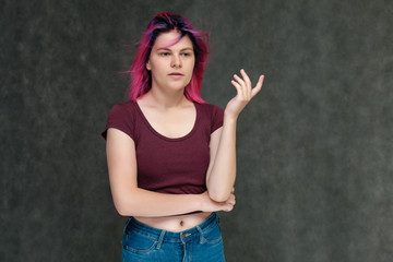 Fototapeta na wymiar Portrait to the waist of a young pretty teen girl in a burgundy T-shirt and jeans with beautiful purple hair on a gray background in the studio. Talking, smiling, showing hands with emotions.