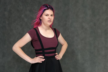 Portrait to the waist of a young pretty teenager girl in a burgundy T-shirt and dress with beautiful purple hair on a gray background in the studio. Talking, smiling, showing hands with emotions.