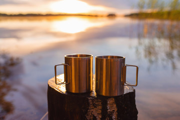 Two stainless steel camping cups or mugs on a wooden stump against a dramatic sunrise or sunshine...