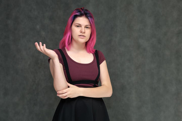 Portrait to the waist of a young pretty teenager girl in a burgundy T-shirt and dress with beautiful purple hair on a gray background in the studio. Talking, smiling, showing hands with emotions.